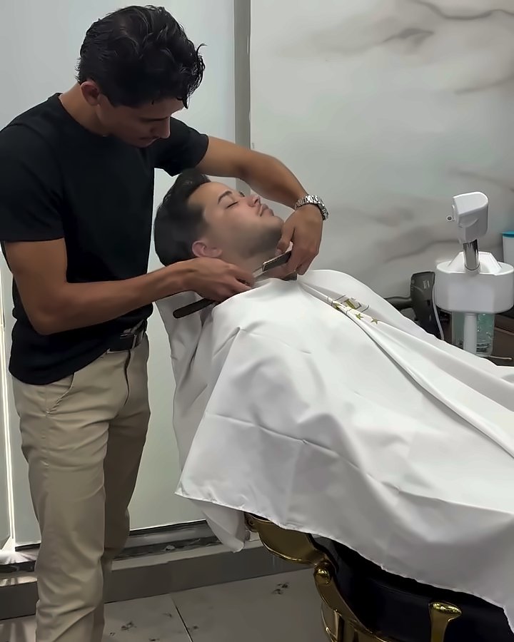 Barbeiro fazendo a barba de um cliente reclinado na cadeira dourada, com a parede de mármore ao fundo