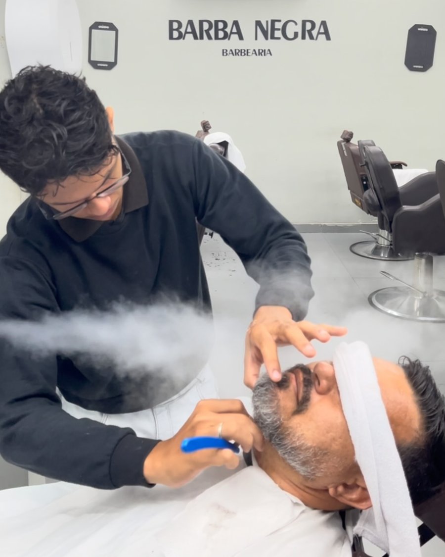 Barbeiro fazendo a barba de um cliente com toalha no rosto e vapor de ozônio, diante do letreiro Barba Negra Barbearia